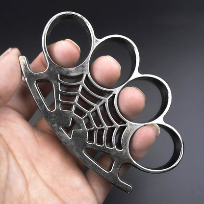 A gray-finish four-finger knuckle duster held in a hand, with four oval finger holes and a spider-web casting with a spider on the palm rest.