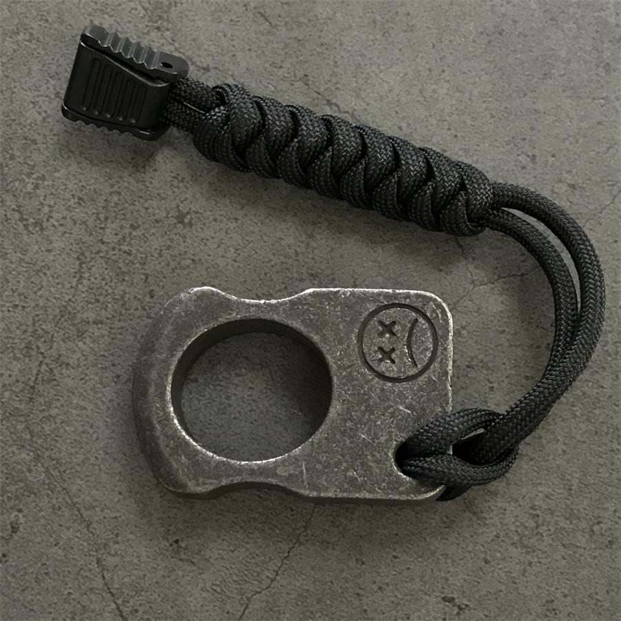 Gray single-hole knuckle weight with black cord lanyard and cord lock laid on a concrete surface