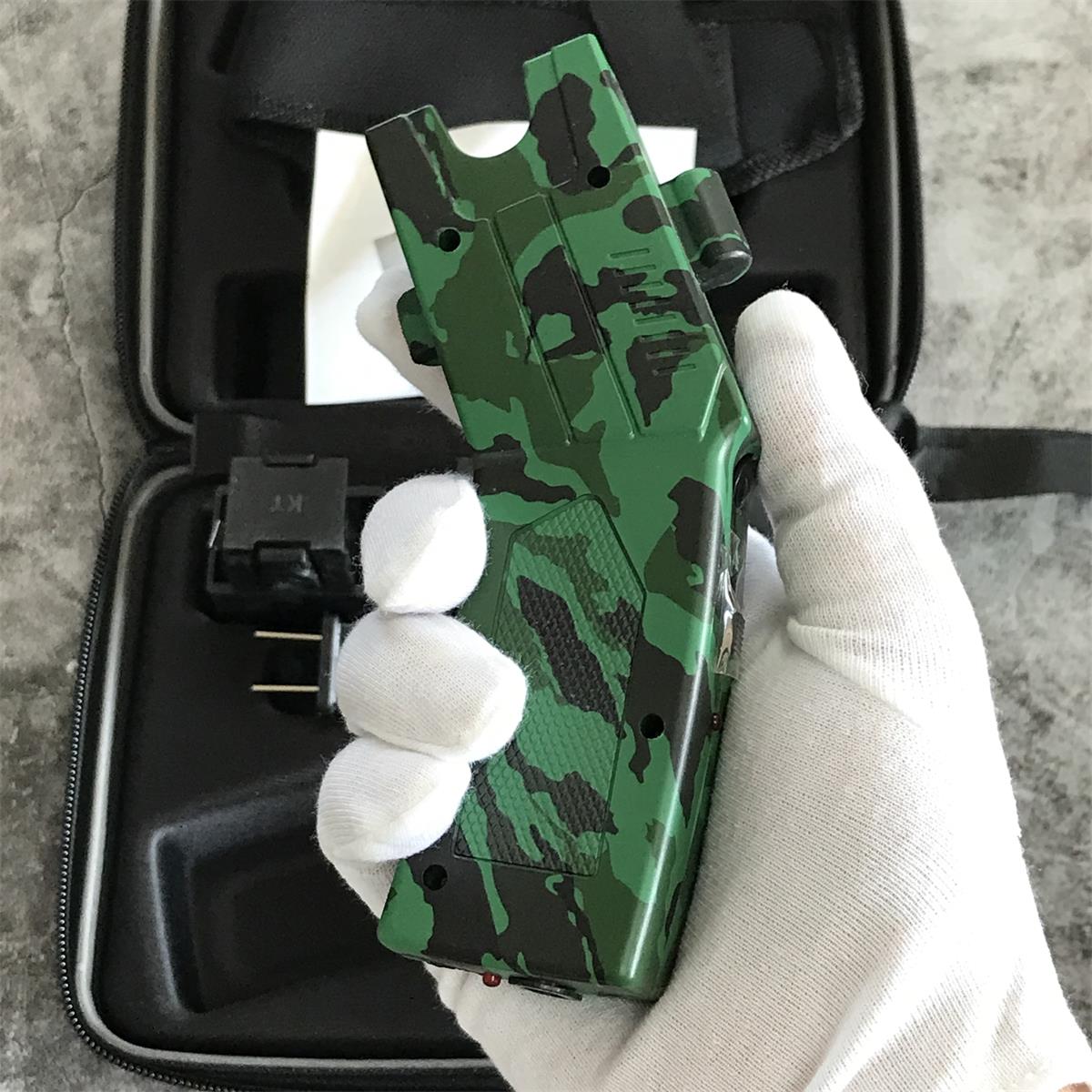White gloved hand holding the green camouflage stun gun unit above the open case showing the top rail, vent slots and red indicator dot