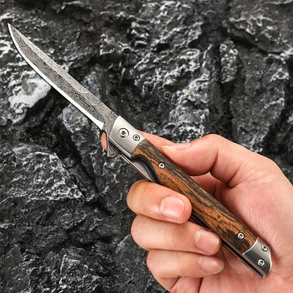 Hand holding an open folding knife with a slim Damascus-pattern drop-point blade and striped wood handle, against dark rock