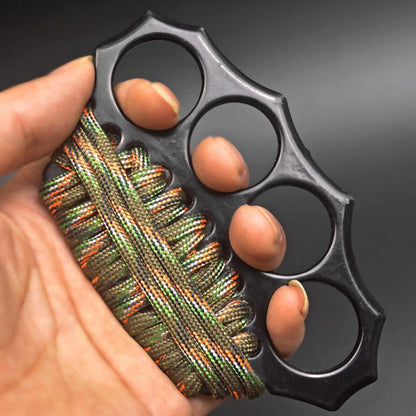 Hand holding the matte black knuckle duster with fingertips through the four holes and a camo paracord wrap on the grip.