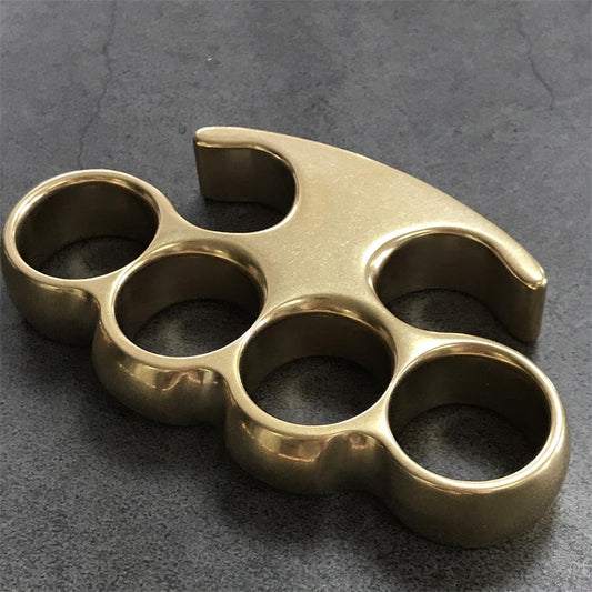 Large brass knuckle duster with claw-tip frame and four round holes, stonewashed finish on a gray concrete surface