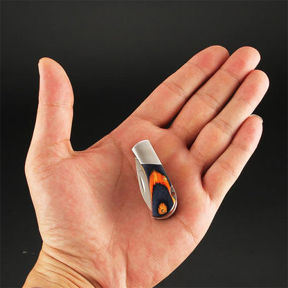 A closed mini folding keychain knife rests in an open palm against a dark background, its navy-and-orange coloured wood handle capped with a brushed silver bolster.