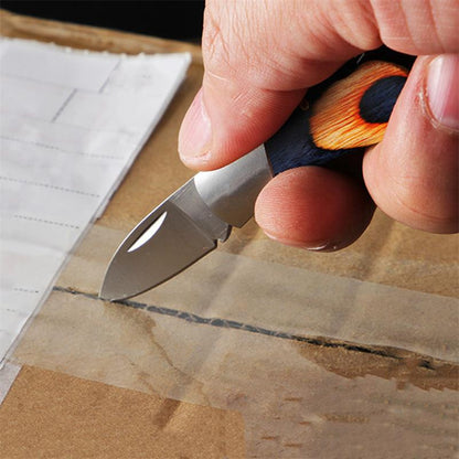 A hand holds the open mini folding knife and slices the clear packing tape along the seam of a cardboard box, with the navy-and-orange wood handle in contrast to the blade.