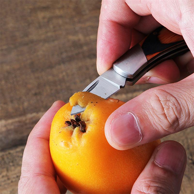 A hand holds the open mini folding knife by its coloured wood handle and cuts into the top of an orange persimmon on a wooden table, showing the silver blade in use.