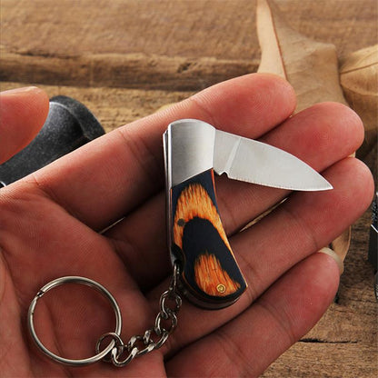 An open mini folding knife held in an open palm, its polished drop-point blade extended from a navy-and-orange wood handle, with a key ring and chain dangling below.