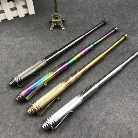 Four pen-style expandable batons in their color variants laid diagonally on a dark textured surface, with a white fence and a bronze Eiffel Tower figurine in the background.