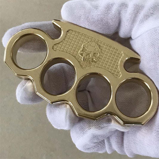 Angled gloved-hand view of the brass mini knuckle duster with the skull panel and curved pointed lower edge in frame