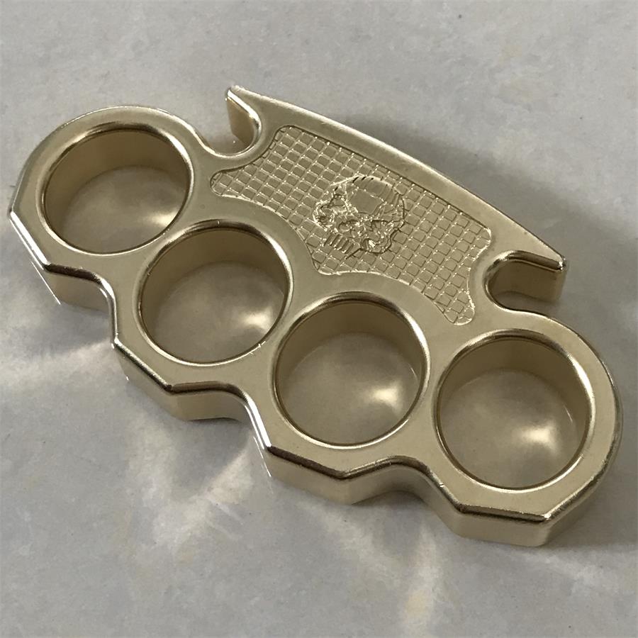 Brass mini knuckle duster lying flat on a light stone surface, top view showing the checkered skull panel