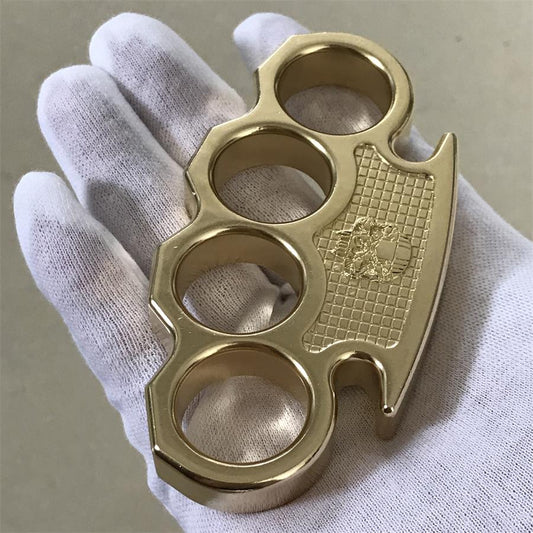 Polished brass mini knuckle duster held in a white-gloved hand, showing the embossed skull checkered panel and four finger holes
