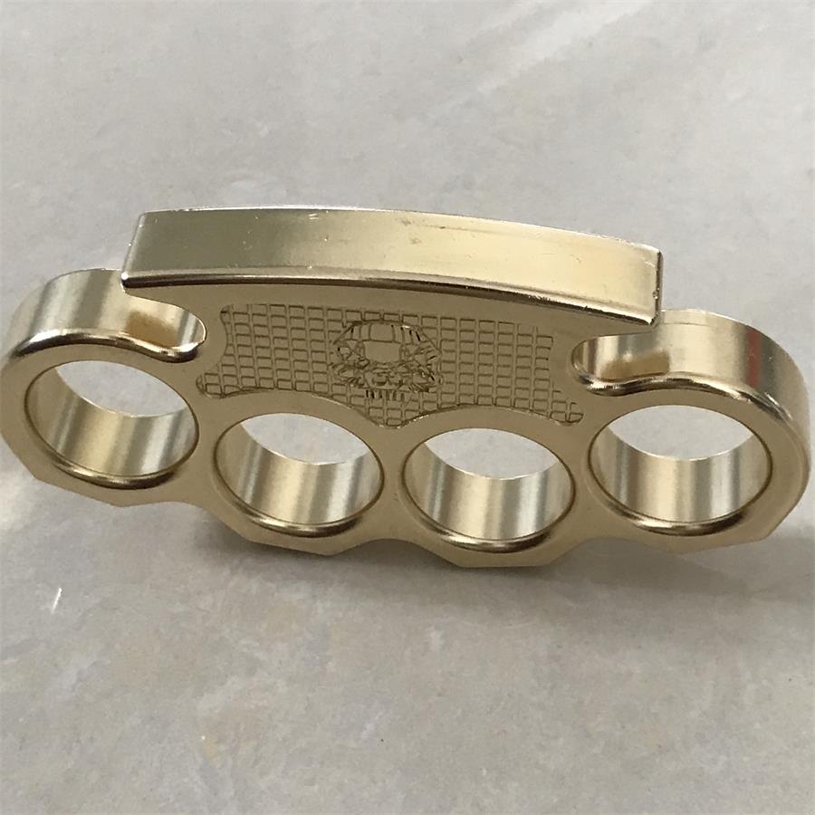 Brass mini knuckle duster standing on a light stone surface, front view with the skull panel centered