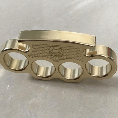 Brass mini knuckle duster standing on a light stone surface, front view with the skull panel centered