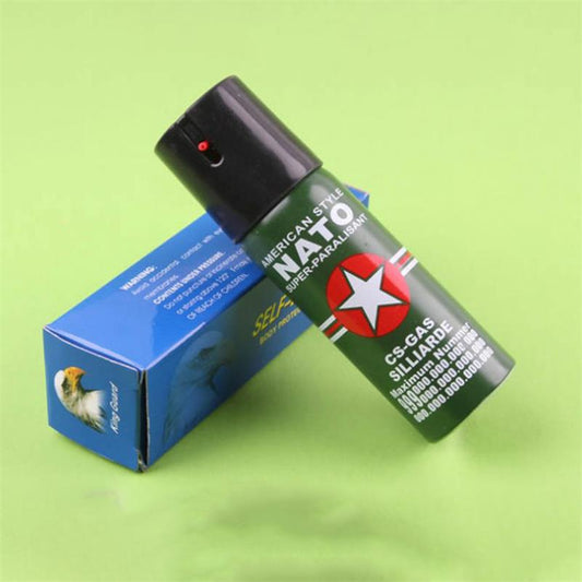 Green NATO-labelled pepper spray canister with black flip-top cap leaning against its blue retail box on a green background