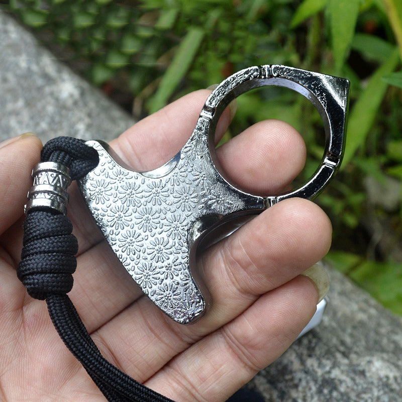 Silver single finger knuckle held in a hand outdoors showing the embossed body, notched finger ring and black paracord lanyard with carved beads