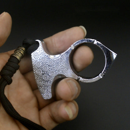 Hand holding the silver one finger knuckle duster with daisy-embossed palm plate, round finger hole and black paracord lanyard on a dark background