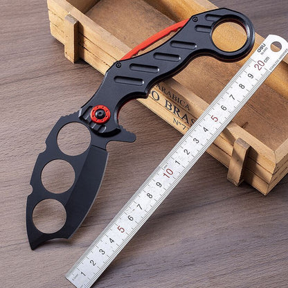 Open black folding knuckle knife with red accents next to a metal ruler on wood