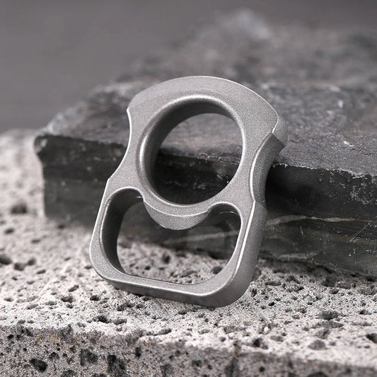 Grey zinc alloy mini knuckle duster bottle opener standing upright on stone with its finger ring facing up