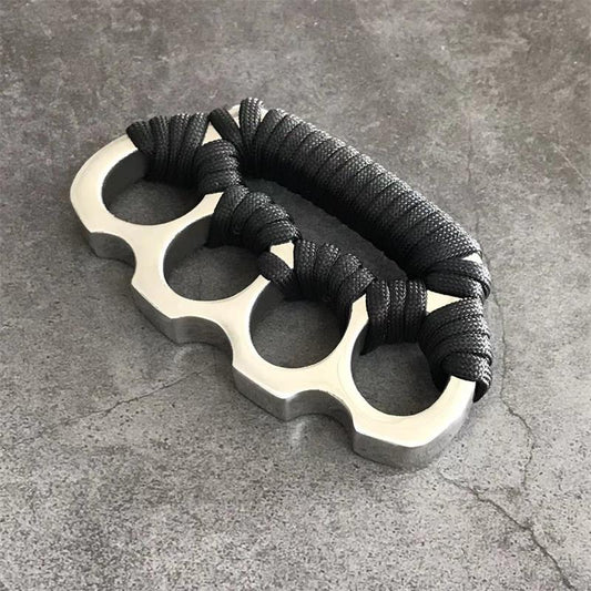Stainless steel parachute rope knuckle duster with black cord wrap lying on gray concrete.