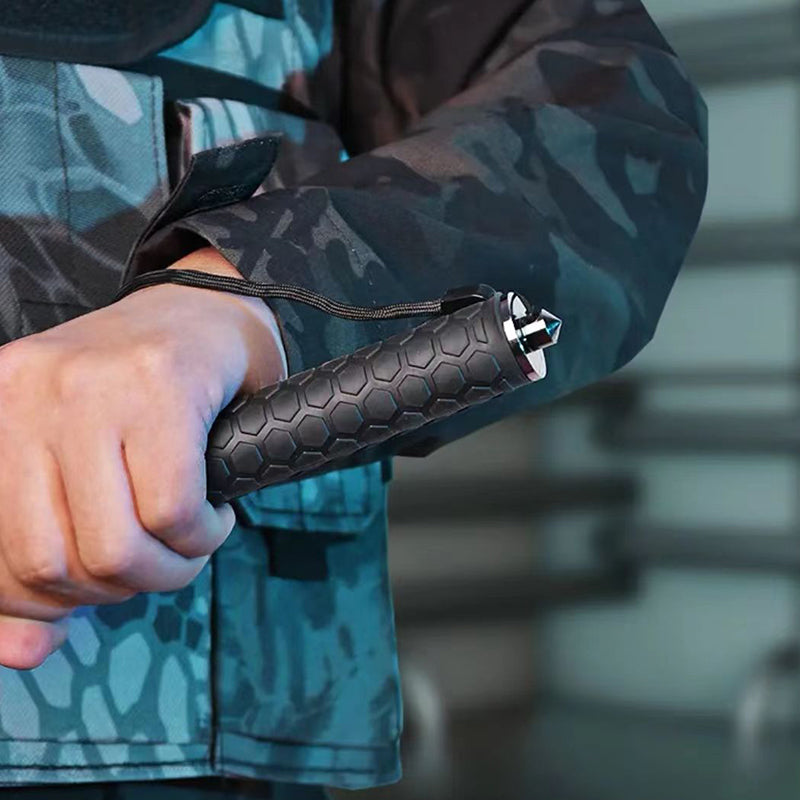 Hand gripping the hexagon-textured closed baton with a paracord lanyard around the wrist and camouflage clothing behind