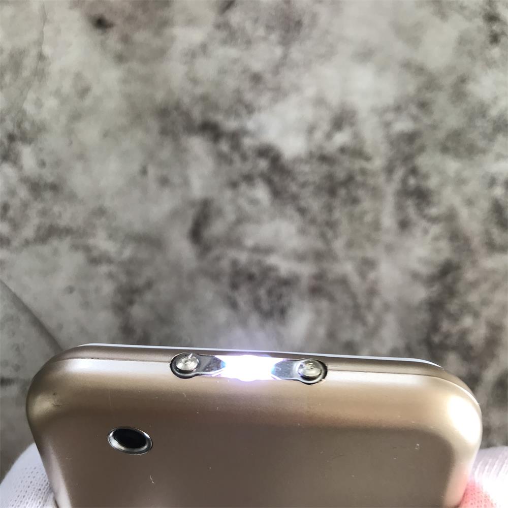 Close-up of gloved fingers holding the gold device with two metal prongs between flanking plates and a bright LED light glowing at the edge