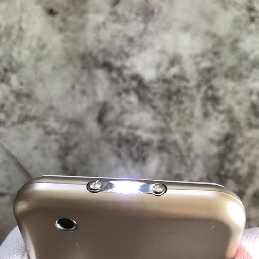 Close-up of gloved fingers holding the gold device with two metal prongs between flanking plates and a bright LED light glowing at the edge