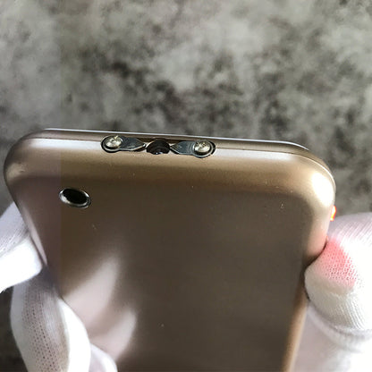 White gloved hand holding the gold phone-shaped device with the twin metal prongs on the short edge and a small round hole on the back