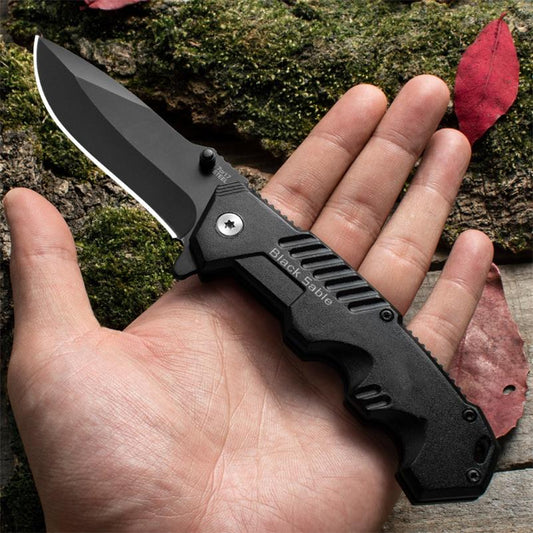 Large black folding knife held in hand over forest moss