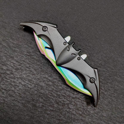 A closed rainbow bat folding knife seen from above on a dark gray surface, the black bat-shaped handle with ears, eyes and wing slots showing iridescent blade edges along both sides.