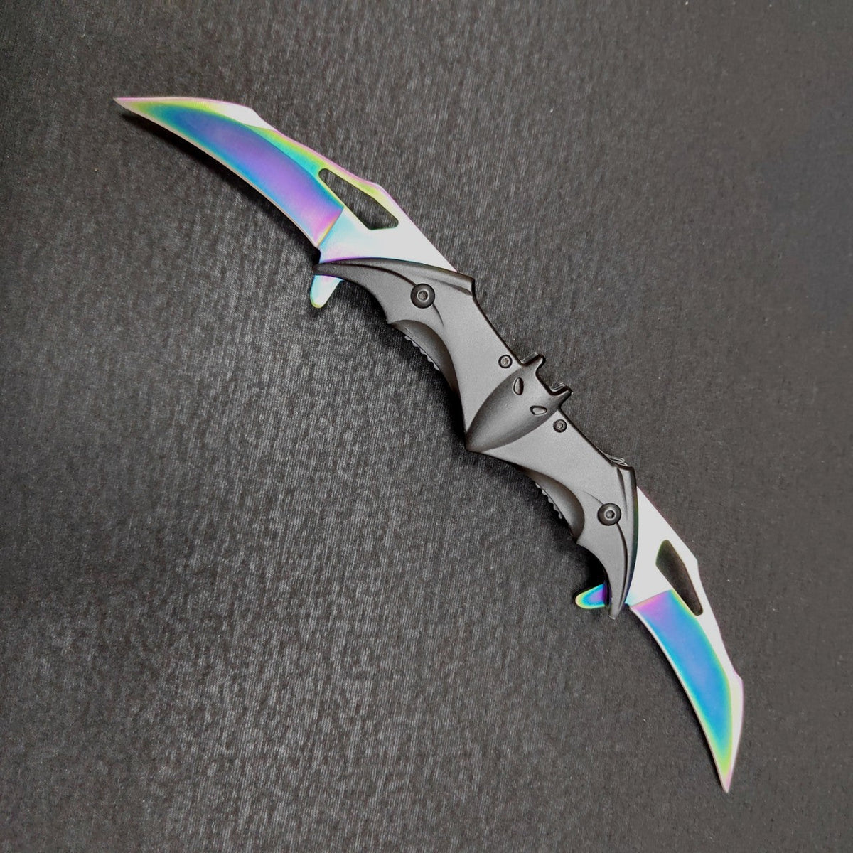 A fully open rainbow-finish bat folding knife lying diagonally on a dark gray surface, two iridescent blades flanking a black bat-shaped body with ears, eyes and slotted wing edges.