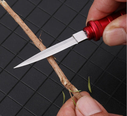 Close-up of the red keychain dagger with the blade out, cutting a wooden stick on a dark grid mat, with a few green sprigs nearby.
