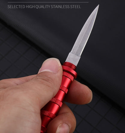 Red keychain dagger held in a hand with the blade open, ribbed handle, on a black grid mat with 'Selected High Quality Stainless Steel' text.