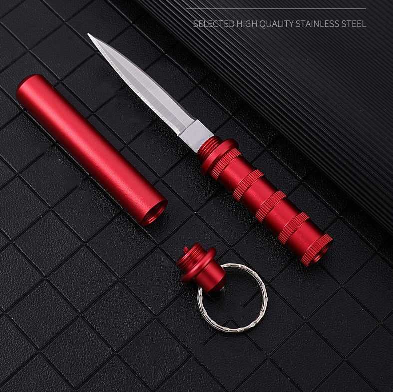 Red keychain dagger laid out with its cylindrical aluminum scabbard and a keyring cap on a black grid mat, with 'Selected High Quality Stainless Steel' text.