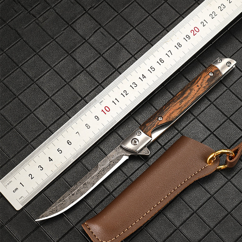 Steel centimeter ruler laid beside an open Damascus-pattern folding knife with a striped wood handle and a brown leather sheath with brass ring, on a dark grid mat