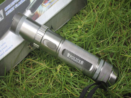 Silver torch-style stun gun on grass showing the printed POLICE badge, ring of head pins and wrist strap in front of its retail box