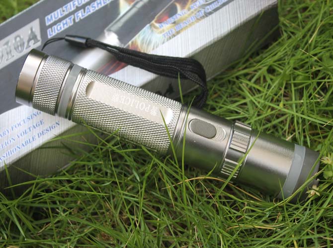 Silver stun gun lying on grass beside its box showing the printed POLICE badge, oval side button and black wrist strap