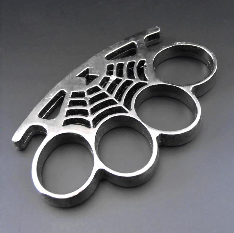 A silver cobweb knuckle duster lying at an angle on a gray surface, showing four oval finger holes and a spider-web casting with a spider on the palm rest.