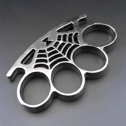 A silver cobweb knuckle duster lying at an angle on a gray surface, showing four oval finger holes and a spider-web casting with a spider on the palm rest.