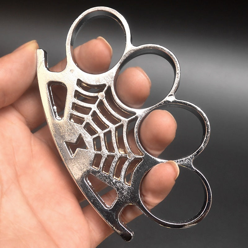 A polished silver four-finger knuckle duster gripped in a hand, its four oval finger holes above a spider-web casting and a small spider on the curved palm rest.