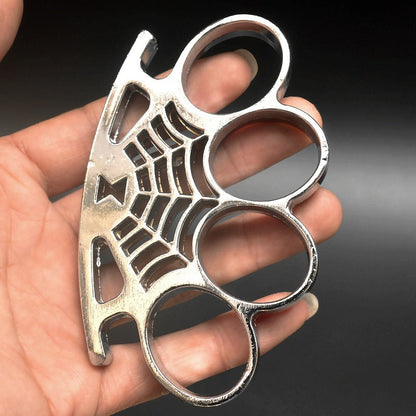 A silver four-finger knuckle duster held in a hand, showing four large oval finger holes and a cast spider-web pattern with a spider on the palm rest.