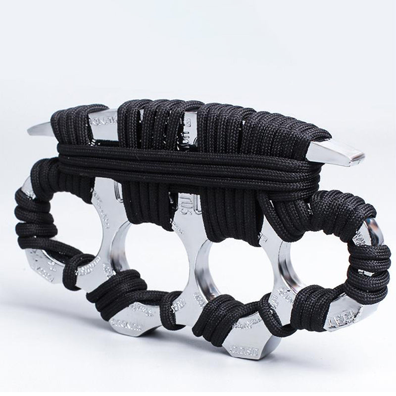 A polished silver zinc alloy knuckle duster wrapped in black paracord, its four finger holes and knuckle faces showing faint engraved lettering, photographed on a plain white background.