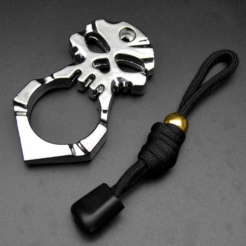 The polished silver version of the skull knuckle duster lying beside its black paracord lanyard on a dark gray surface, showing the eye sockets, heart-shaped hole and teeth cut-outs above the single finger ring.
