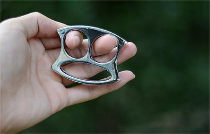Silver two-hole pocket EDC knuckle duster held in a bare hand against a green outdoor background.