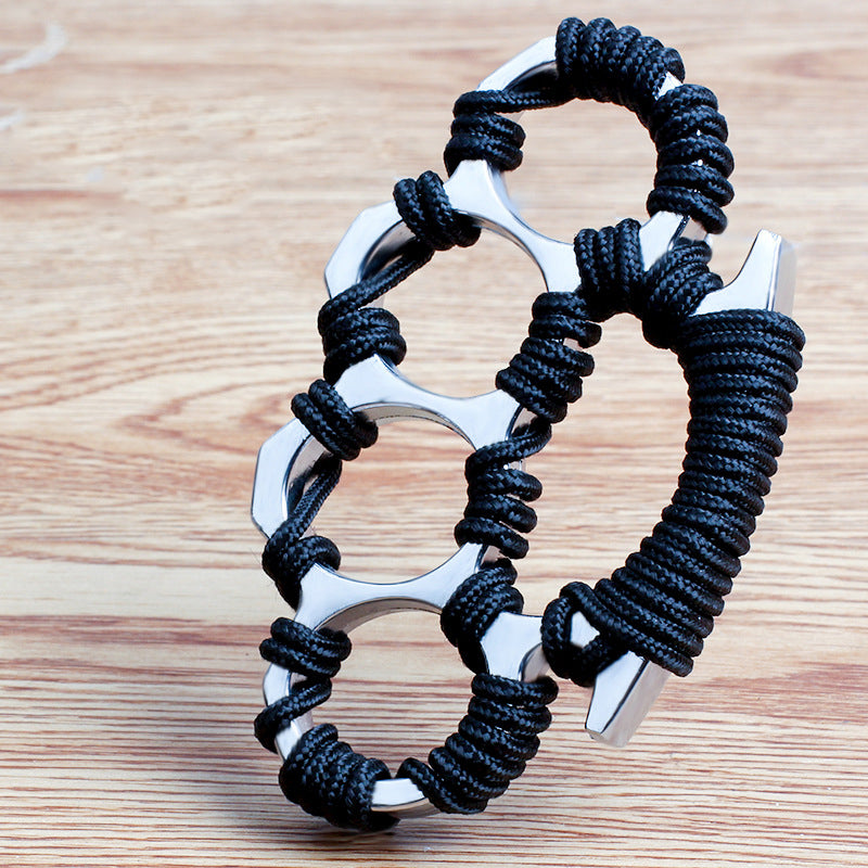 Angled view of the silver woven knuckle duster with black umbrella rope around the four openings and wrapped palm base on a wooden table