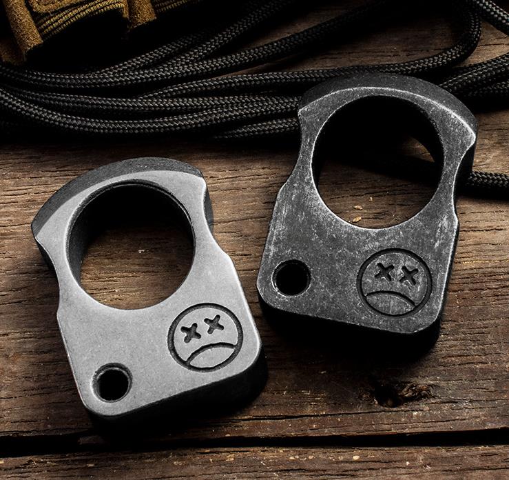 Two single-hole steel knuckle weights with crossed-eye face engravings, one light and one dark, on a wooden surface with coiled black paracord behind