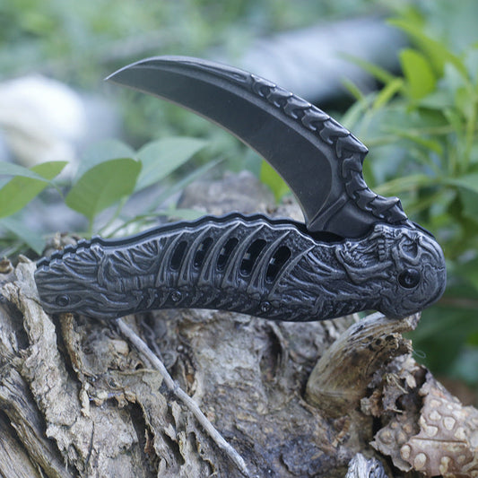 An open folding knife rests on rough tree bark outdoors, its curved stone-washed hawksbill blade above a dark sculpted handle detailed with a skull at the pivot end and a ribbed, spine-like channel along the body.