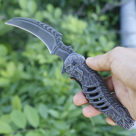 A hand holds an open folding knife against blurred green foliage, showing a dark stone-washed curved hawksbill blade with an embossed spine pattern and a sculpted gray handle shaped like a skull with rib-like cutouts.