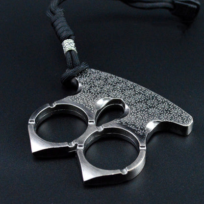 Steel gray two-finger knuckle with floral engraving lying flat on a dark surface, cord through the top