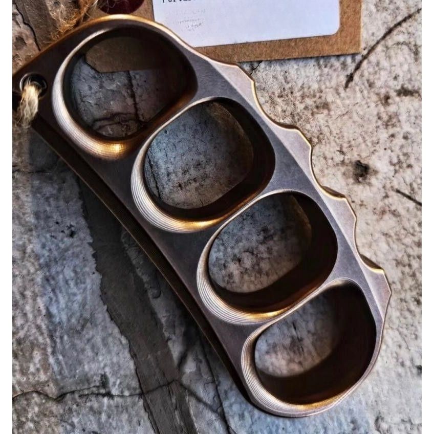 Bronze toned steel knuckle hanging by a natural twine cord through its lanyard hole above a kraft paper tag