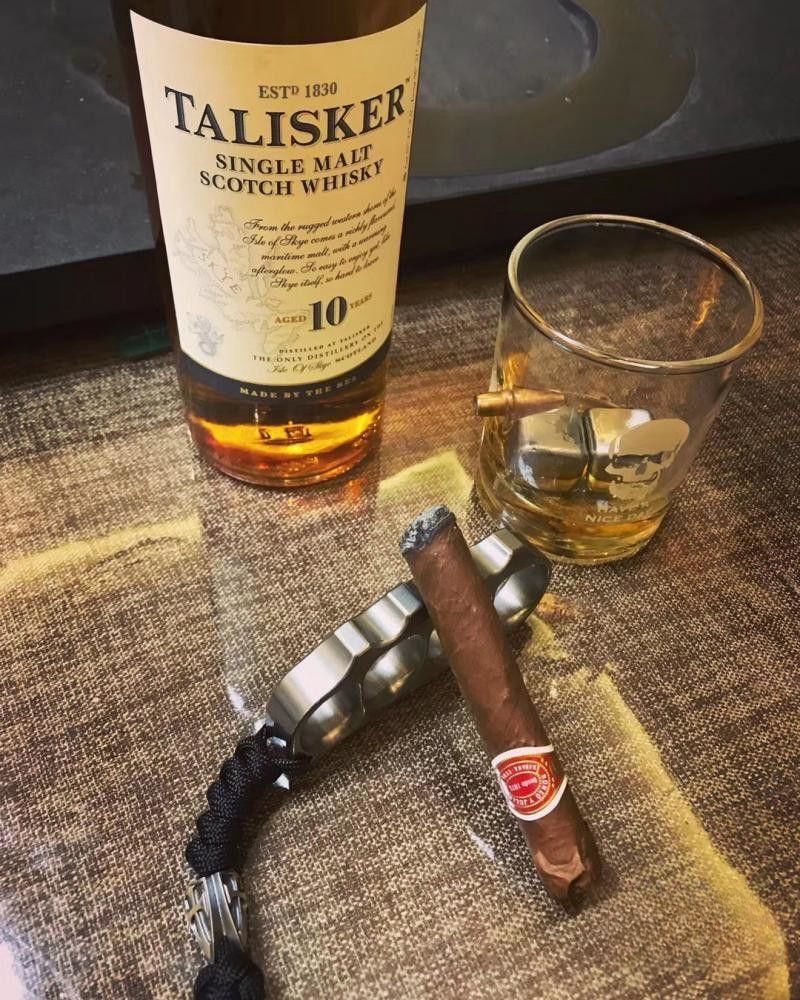 Polished steel knuckle with a black paracord lanyard lying on a table beside a whisky bottle and a cigar