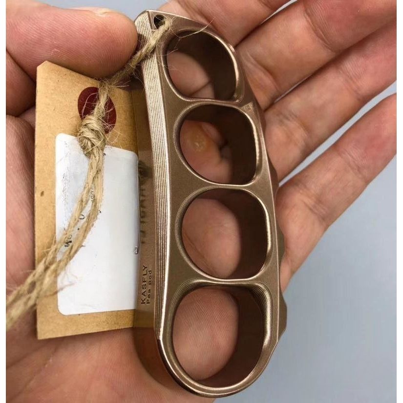 Hand holding the bronze toned steel knuckle with a kraft paper tag tied through the lanyard hole